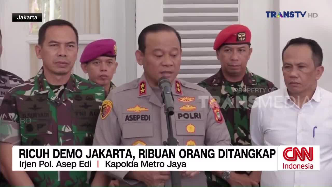A police officer in uniform speaks into a microphone, flanked by men in military attire and civilian clothes, all standing in a room. The news is being broadcast on Trans TV from Jakarta, Indonesia.
