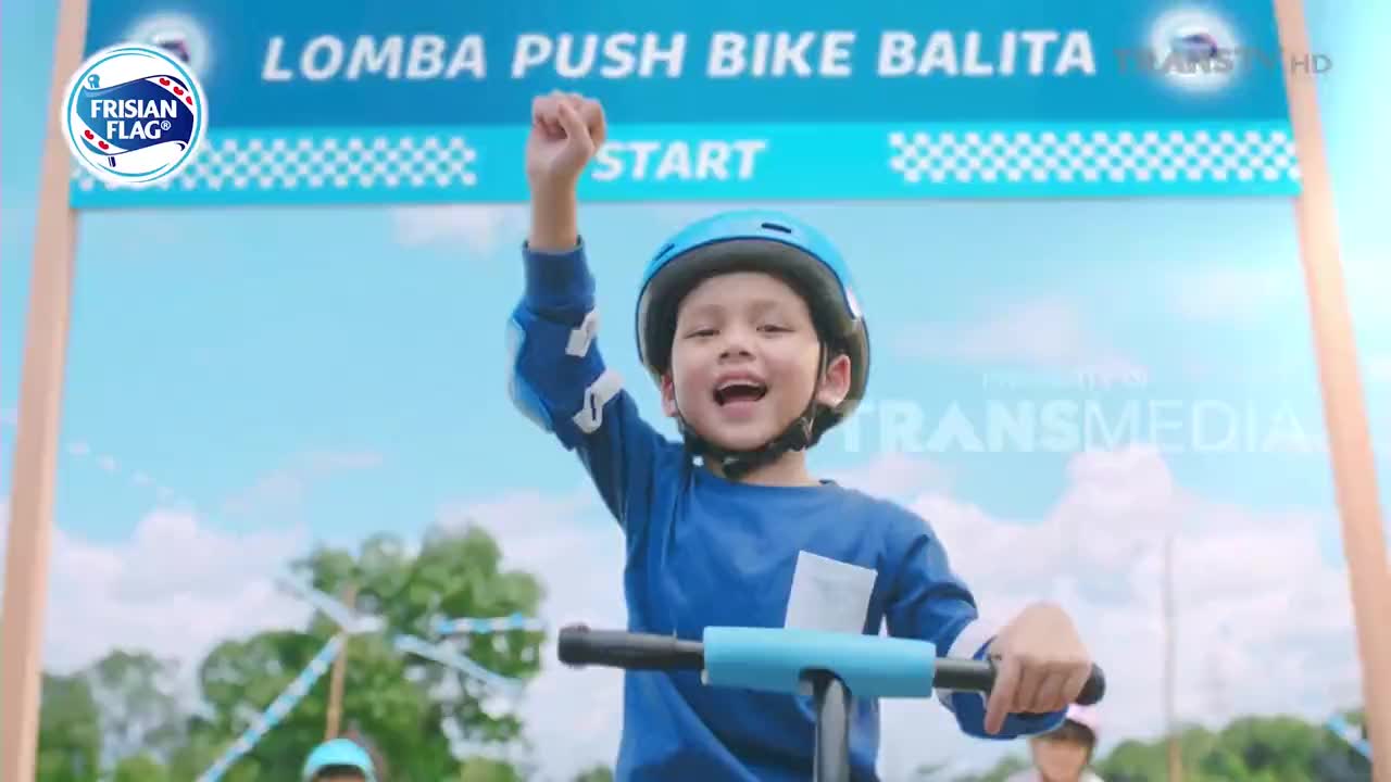 A young boy on a push bike, wearing a helmet, raises his fist in victory as he crosses the finish line. The scene is part of a "Lomba Push Bike Balita" race, as broadcast by Trans TV in Indonesia.
