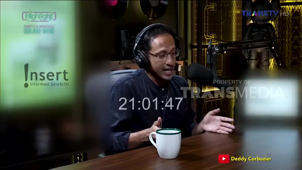 A man wearing glasses and headphones gestures with his hands while speaking into a microphone, presumably on a Trans TV show. A mug sits on the table in front of him, and the time is displayed on the screen.
