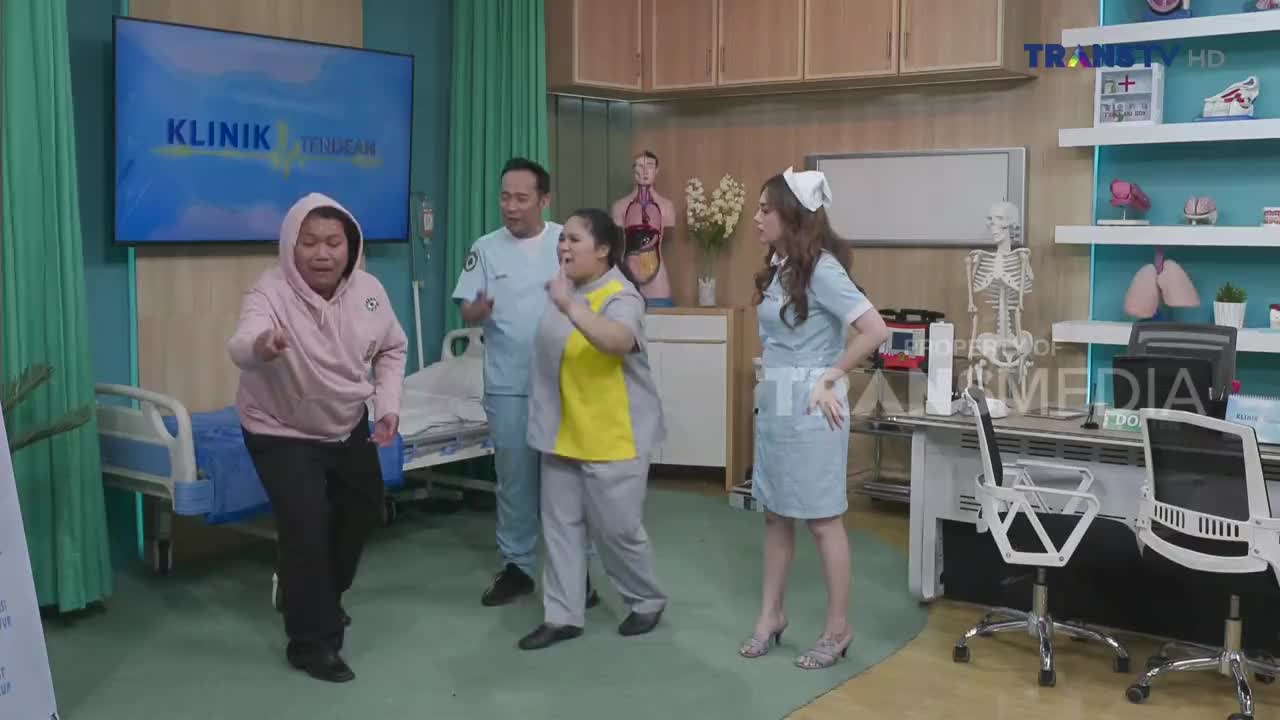 On the set of what looks like a Trans TV show, a group of people are dancing in a mock-up hospital room. The person in the pink hoodie is pointing and moving their feet, while the others are doing a similar dance.
