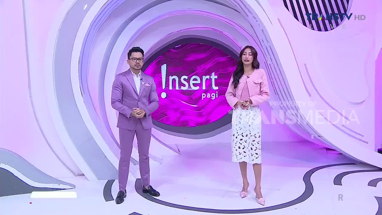 Two presenters, a man in a lavender suit and a woman in a pink jacket, stand in front of a backdrop for "Insert Pagi" on Trans TV. The set design features swirling, pastel-colored shapes.
