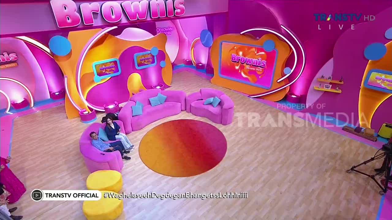 The set of Brownis on Trans TV is awash in pink, with a couple of people sitting on a curved sofa. The show's logo is displayed on multiple screens throughout the studio.
