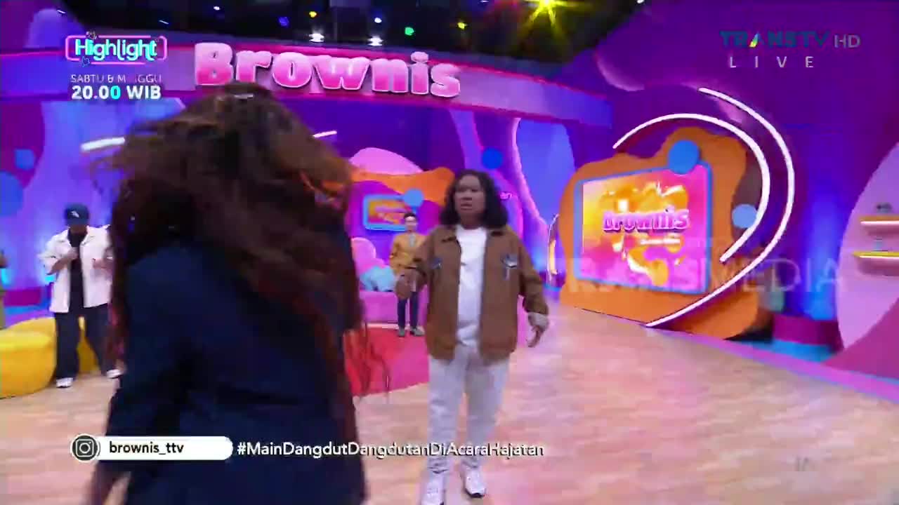 A woman's hair whips around as she moves toward the camera, obscuring the view of a man in a brown jacket.  He appears to be walking toward her on the brightly lit set of "Brownis" on Trans TV from Indonesia.
