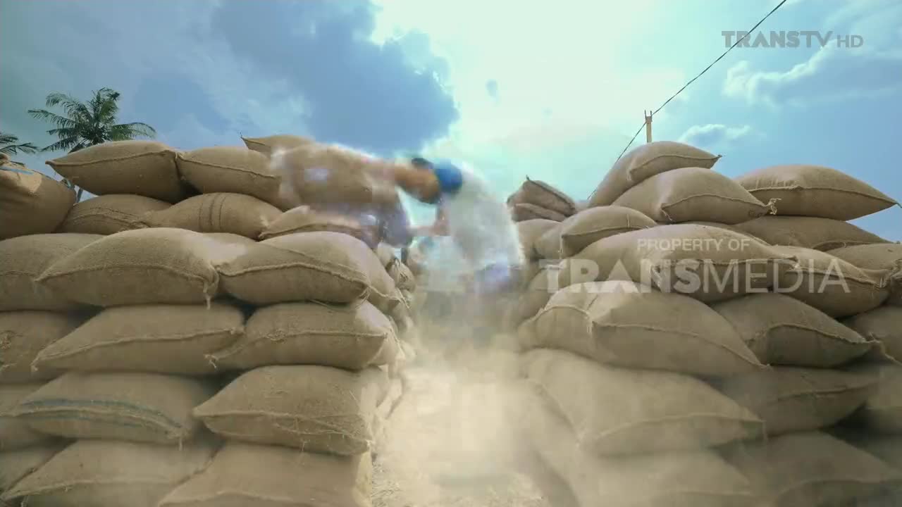 A worker, obscured by a cloud of dust, is moving through a narrow passage between towering stacks of burlap sacks. The scene is under a bright, Indonesian sky, and the Trans TV logo is visible in the corner.
