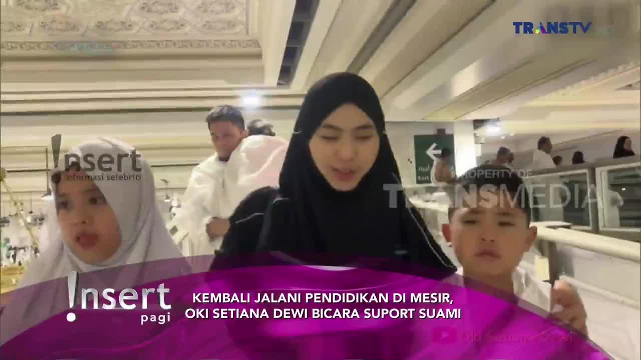A woman in a black hijab speaks directly to the camera, flanked by two children, all in white. Behind them, other people in white robes walk through a bright, ornate space, likely a religious site.
