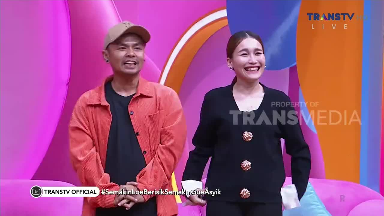 The two people on the Trans TV stage are smiling, the woman in a black blazer with gold buttons, and the man in a red corduroy shirt. They're standing in front of a pink backdrop, ready for the show.
