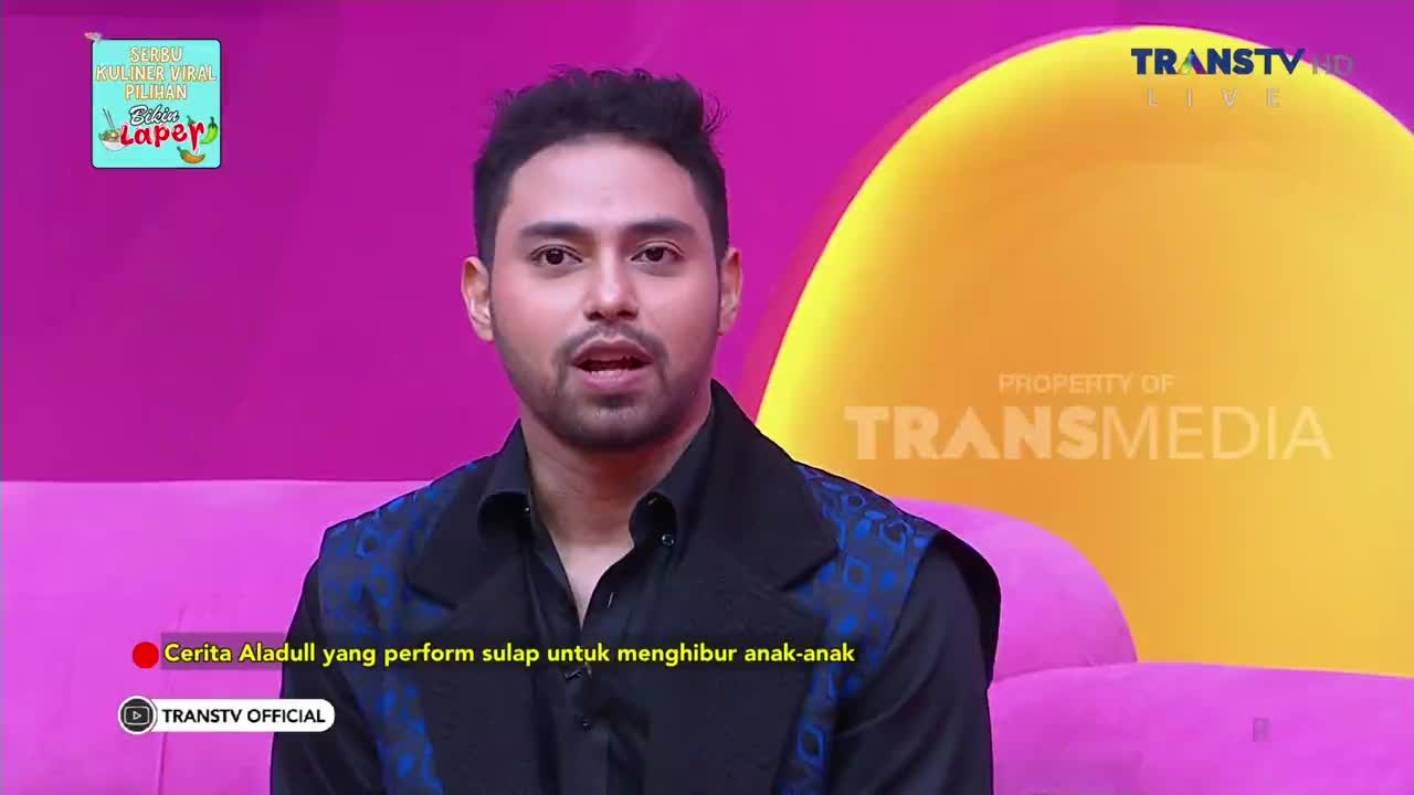 A man in a dark suit is speaking on a Trans TV set, his words likely about a magic performance for children. The background is a mix of bright pink and yellow, with the Transmedia logo visible.
