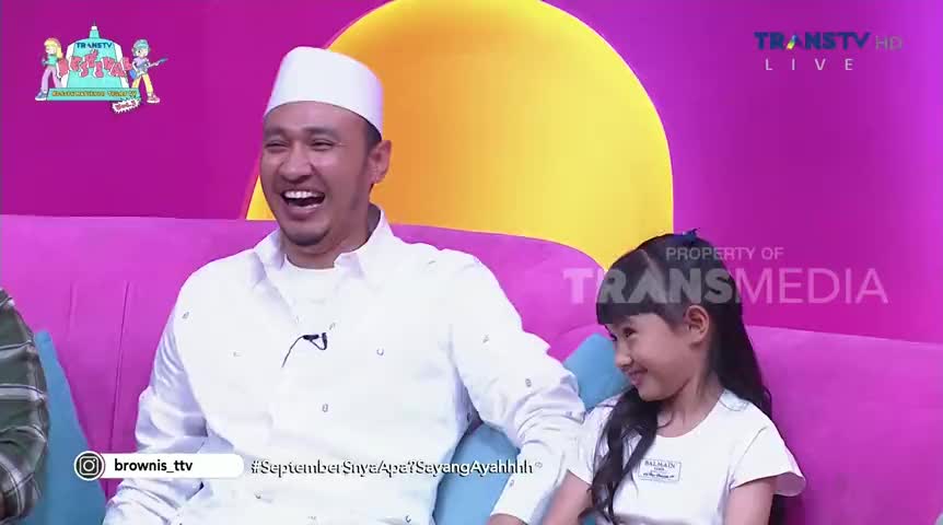 A man in a white cap and shirt is laughing heartily, his face lit up. Beside him, a young girl in a white shirt smiles, looking towards him on the set of an Indonesian Trans TV show.
