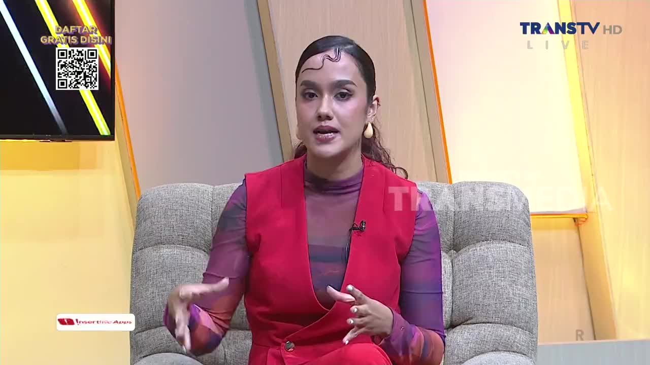 A woman in a red vest gestures with her hands as she speaks, seated on a gray couch. The Trans TV logo is visible on her vest and in the corner of the screen.
