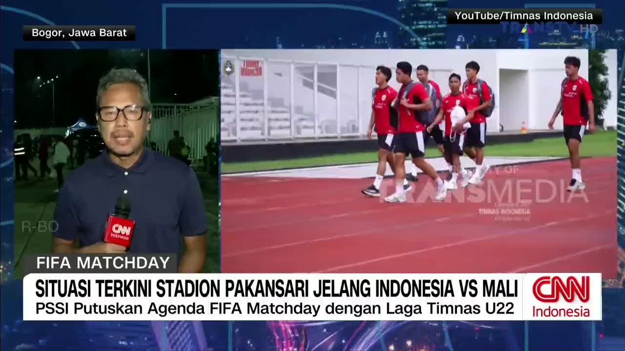 A group of soccer players in red shirts and black shorts walks along a red track. A CNN Indonesia reporter is speaking on camera, with the stadium in the background.
