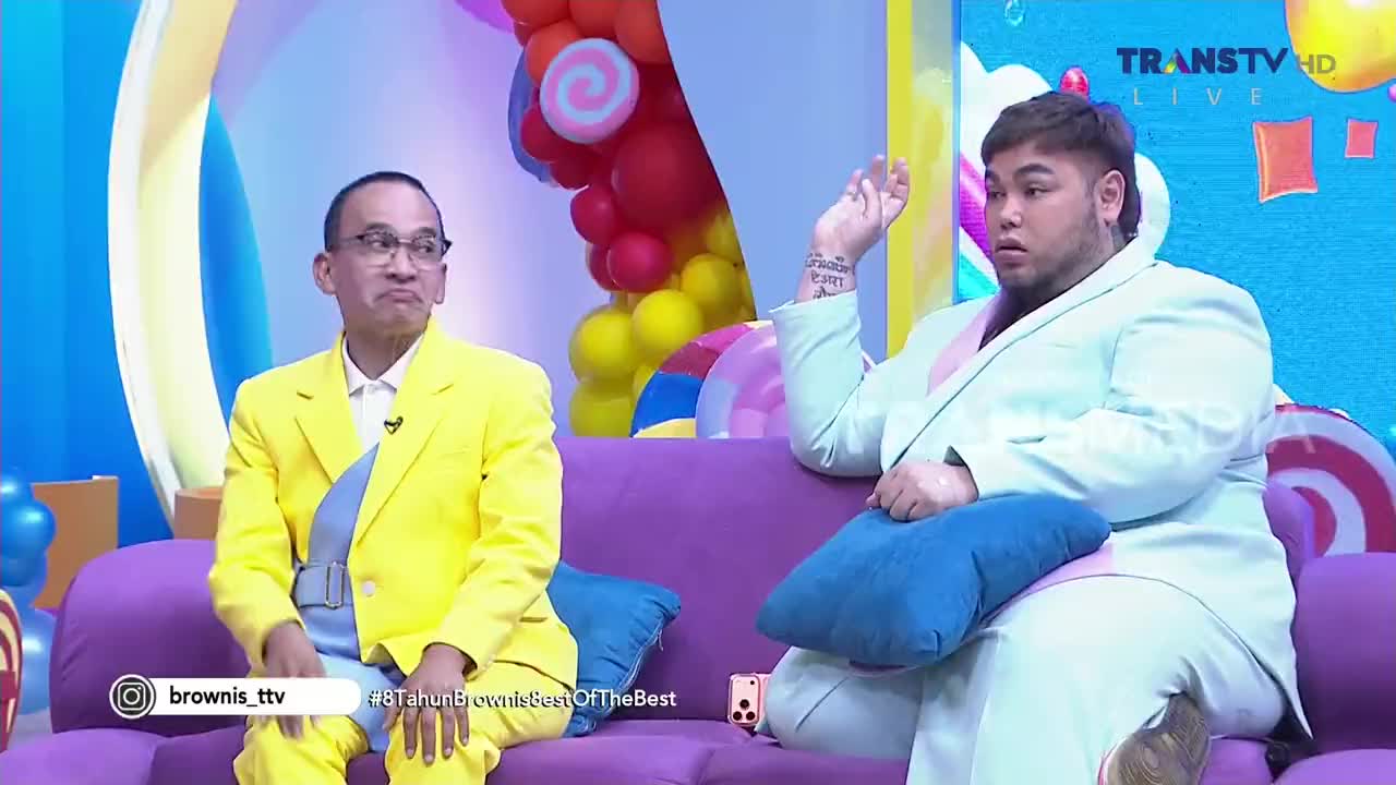 On a Trans TV set in Indonesia, a man in a bright yellow suit sits with a serious expression, while the other, in a pale green suit, gestures with his hand as if making a point. The scene is brightly lit with colorful decorations in the background, likely a studio for a show.
