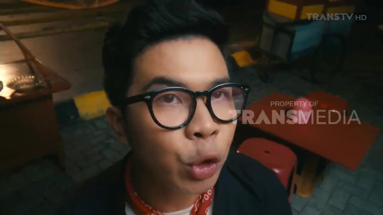 The man with glasses is looking directly at the camera, his mouth slightly open as if he's about to speak. The scene looks like it's from a Trans TV show, filmed in Indonesia.
