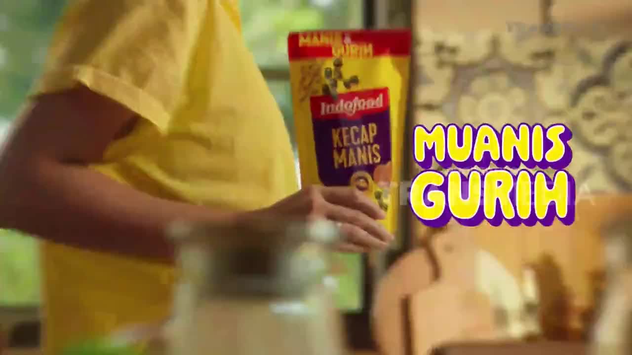 A person in a yellow shirt holds a package of Indofood Kecap Manis, the brand prominent on the packaging. The words "Muanis Gurih" are displayed in a cartoonish font, likely part of an Indonesian television commercial.
