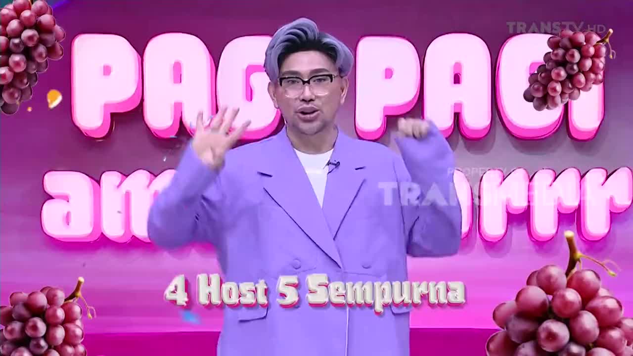 A man in a purple suit gestures with both hands in front of a bright pink backdrop. The Trans TV logo is visible as he speaks on the Indonesian television program.
