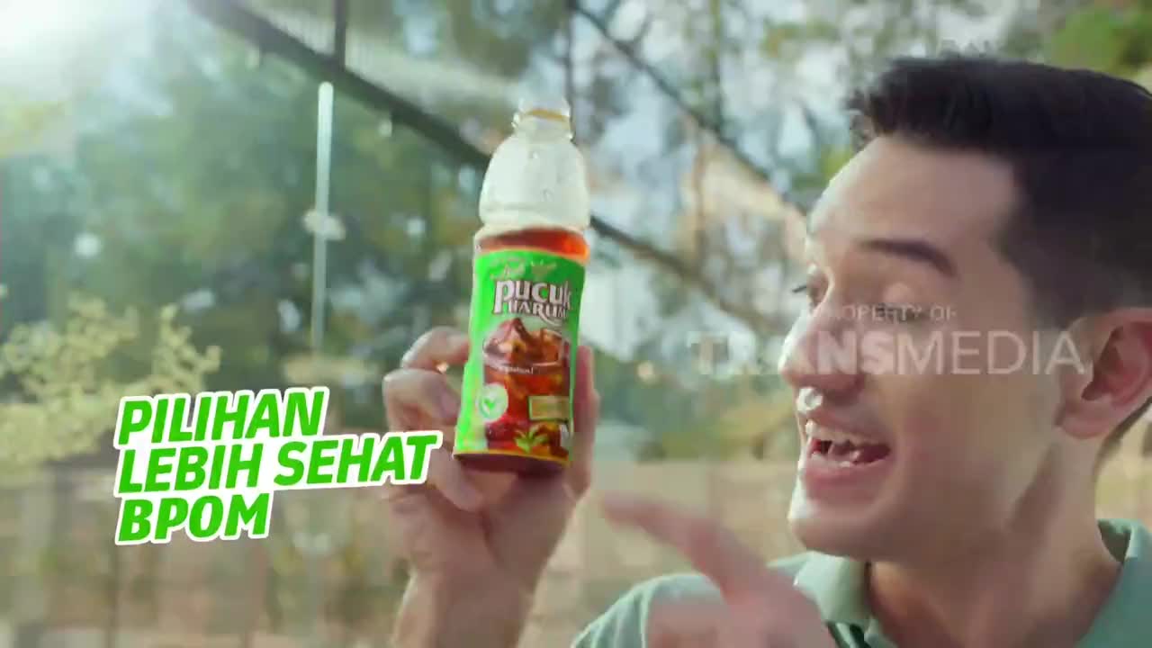 A man holds up a bottle of tea, pointing at it with enthusiasm while smiling. The scene is likely a commercial, as indicated by the text and logo from Trans TV.
