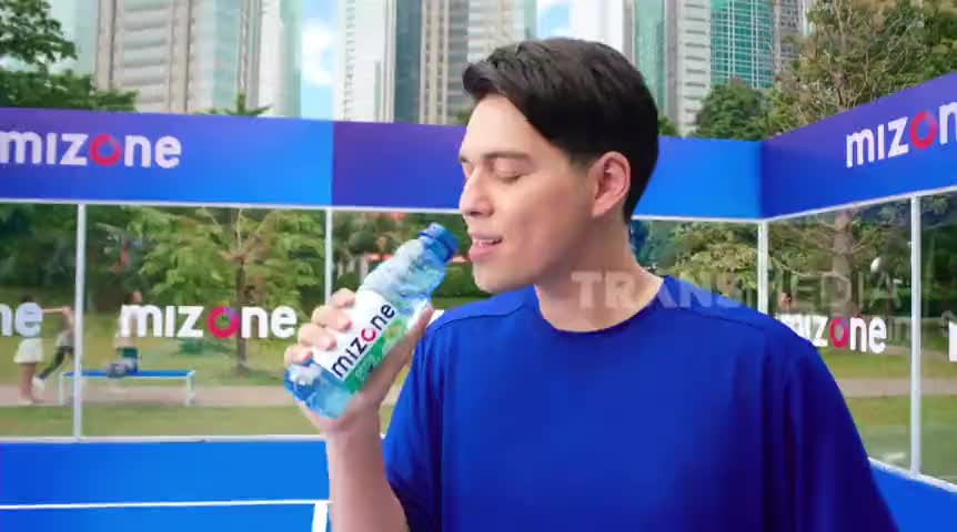 A man in a blue shirt is tilting his head back, taking a drink from a "mizone" bottle. The scene unfolds on what looks like a set for Trans TV, with a park backdrop visible behind him.
A man in a blue shirt is tilting his head back, taking a drink from a "mizone" bottle. The scene unfolds on what looks like a set for Trans TV, with a park backdrop visible behind him.