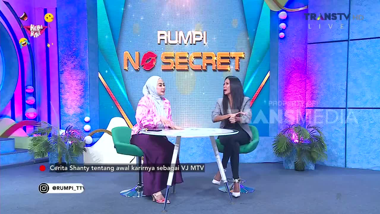 Two women sit across from each other at a white table on the set of "Rumpi No Secret," a Trans TV program. The woman on the left wears a pink hijab and a purple skirt, while the woman on the right has on a grey blazer and black pants. Two women sit across from each other at a white table on the set of "Rumpi No Secret," a Trans TV program. The woman on the left wears a pink hijab and a purple skirt, while the woman on the right has on a grey blazer and black pants.