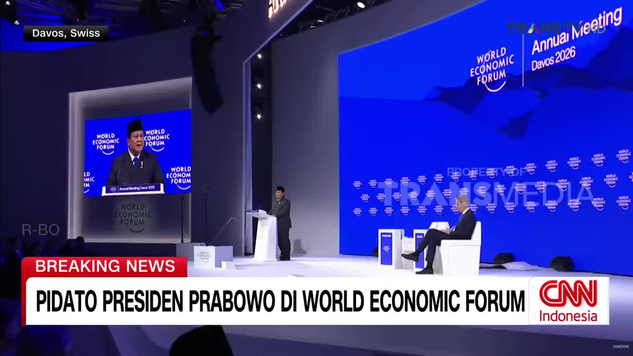 President Prabowo is speaking from a podium on a large screen, while another man stands at a smaller podium on stage. A man sits in a white chair to the right, facing the stage. President Prabowo is speaking from a podium on a large screen, while another man stands at a smaller podium on stage. A man sits in a white chair to the right, facing the stage.