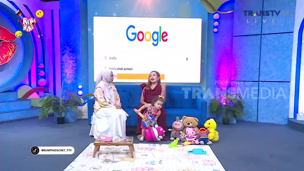 A young girl in a colorful dress plays with toys on a mat in front of a couch. Two women sit on the couch, one in a hijab and the other in a maroon dress, while a large screen behind them displays a Google search bar. A young girl in a colorful dress plays with toys on a mat in front of a couch. Two women sit on the couch, one in a hijab and the other in a maroon dress, while a large screen behind them displays a Google search bar.