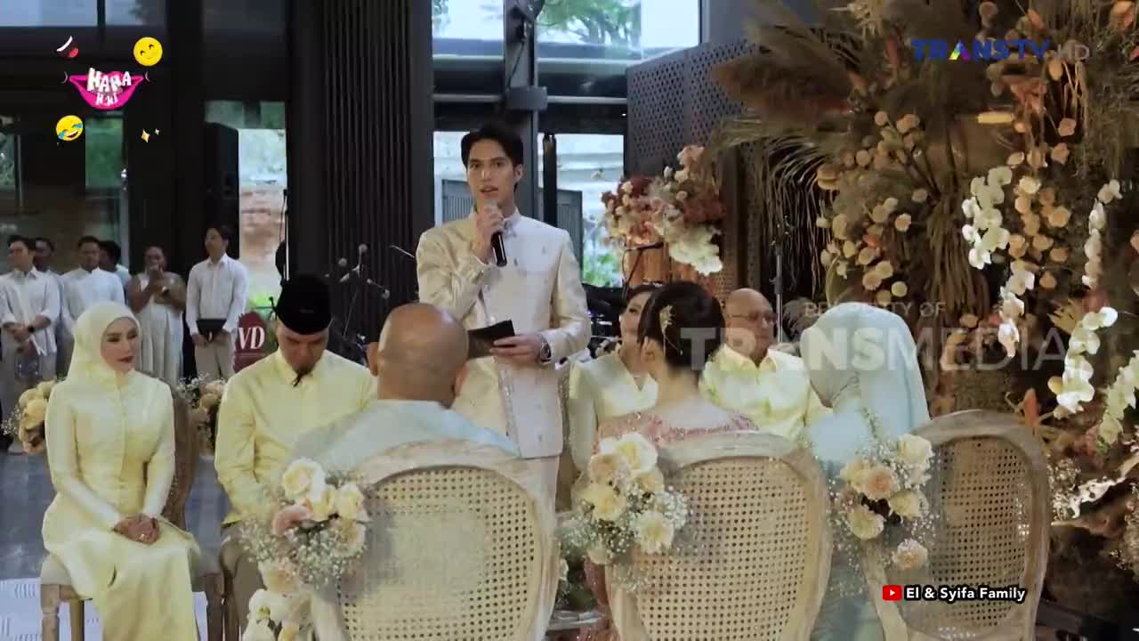 A young man in a cream suit holds a microphone and speaks into it. The bride and groom, dressed in traditional attire, sit facing him.