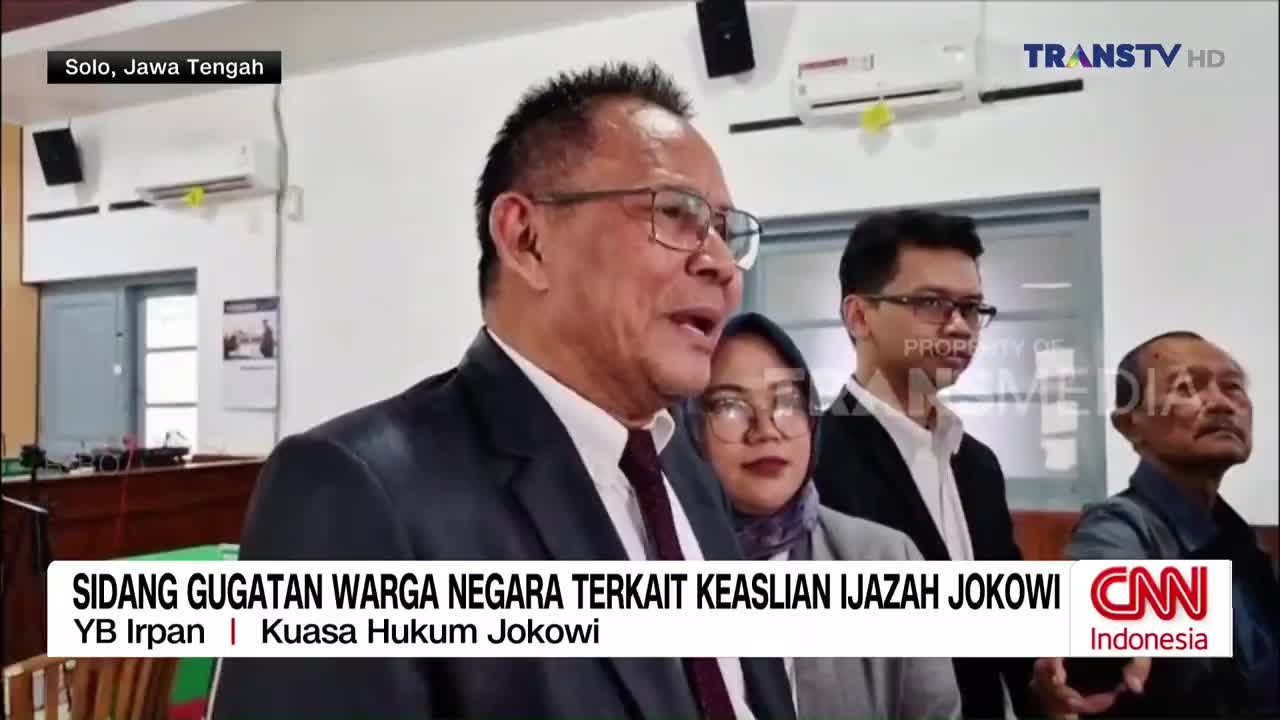 A man in a suit and glasses speaks animatedly, his mouth open mid-sentence. Behind him, a woman in a hijab and another man in glasses listen intently, with a third man looking up and to the side. The Trans TV logo is visible in the upper right corner.