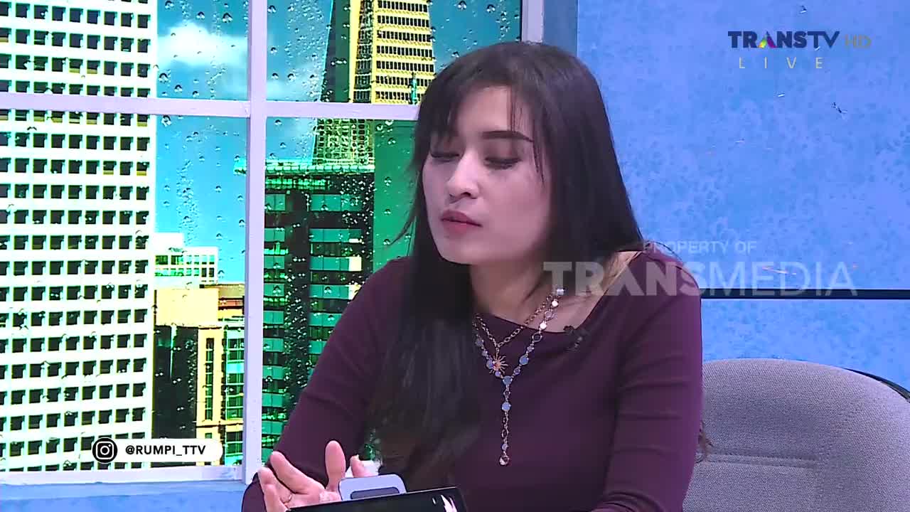 A woman with dark hair and layered necklaces looks down at something in her hands. Behind her, a window shows a city skyline with raindrops streaking down the glass, a common sight during Jakarta's rainy season. The Trans TV logo is visible on the screen.