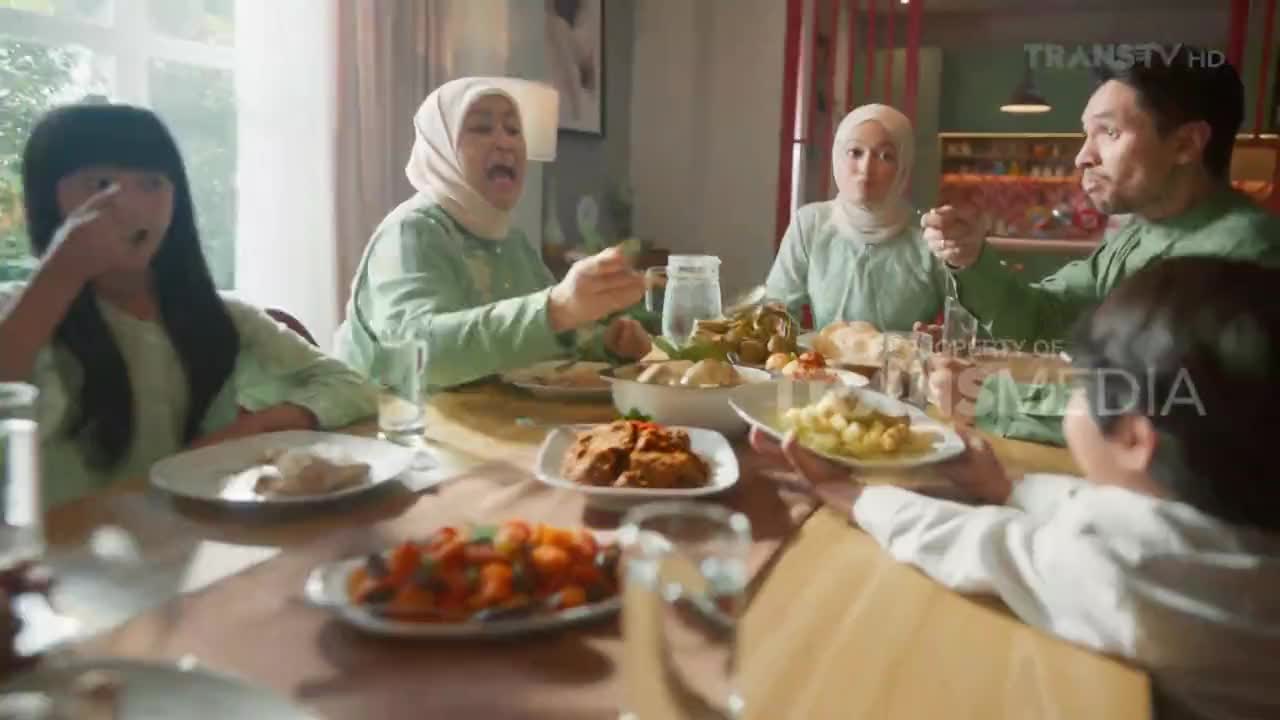 A family gathers around a table laden with Indonesian dishes. The grandmother laughs heartily as she gestures with her fork, while a young girl to her left brings food to her mouth. The Trans TV logo is visible in the upper right corner.