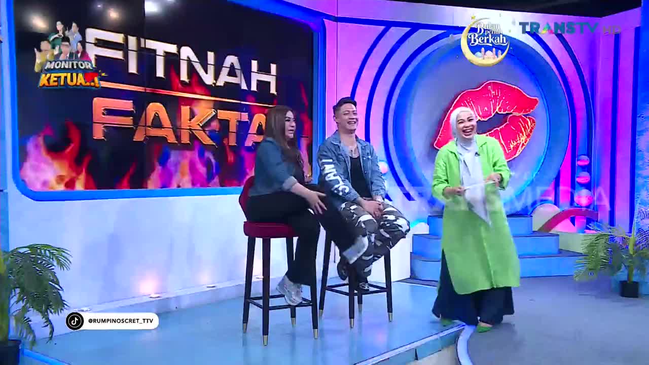 A woman in a bright green jacket laughs as she holds a piece of paper, standing on a stage with a large red lip graphic behind her. Two guests, a man and a woman, sit on stools in front of a screen displaying "FITNAH FAKTA" with fiery graphics, likely for a Trans TV program.