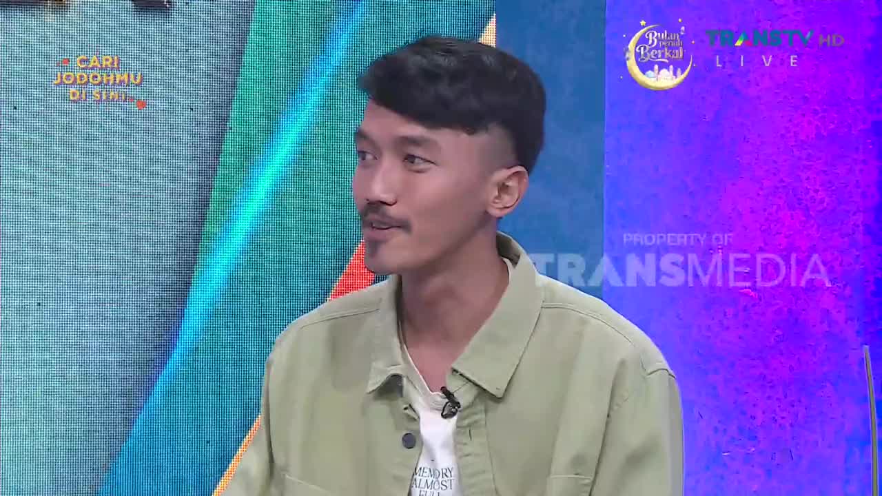 A young man with a mustache and styled hair is on a Trans TV set, wearing a light green jacket over a white t-shirt. The background displays a vibrant, abstract design with text in Indonesian.