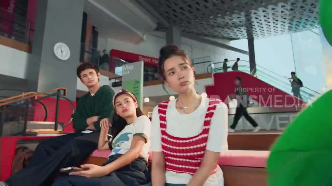 Three young people sit on a bench, a boy in a green hoodie and two girls, one with a headband. In the background, people walk up an escalator in what looks like a modern Indonesian mall.