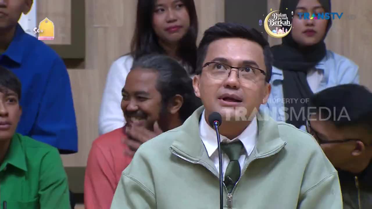 A man in a light green jacket speaks into a microphone, his gaze directed upwards. Behind him, a man in a coral shirt laughs, his hand near his mouth.