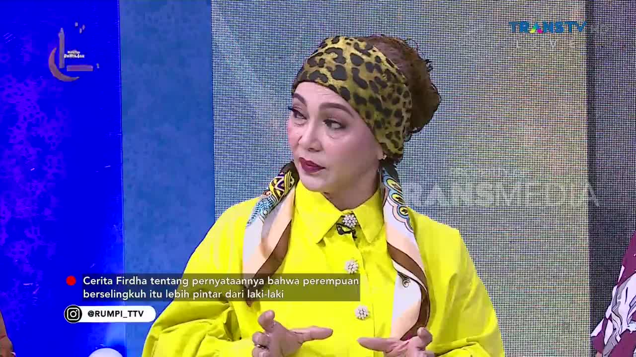 A woman in a bright yellow shirt and leopard-print headscarf speaks animatedly on a Trans TV set. Her hands gesture as she talks, her expression engaged.