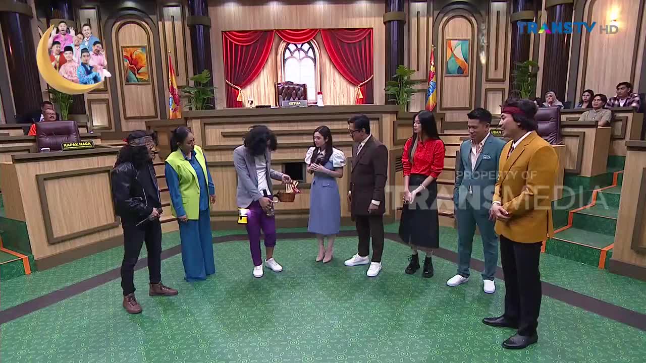 A group of people stands on a green stage, some in colorful outfits. One man in purple pants bends down, holding a small basket. The Trans TV logo is visible on the screen.