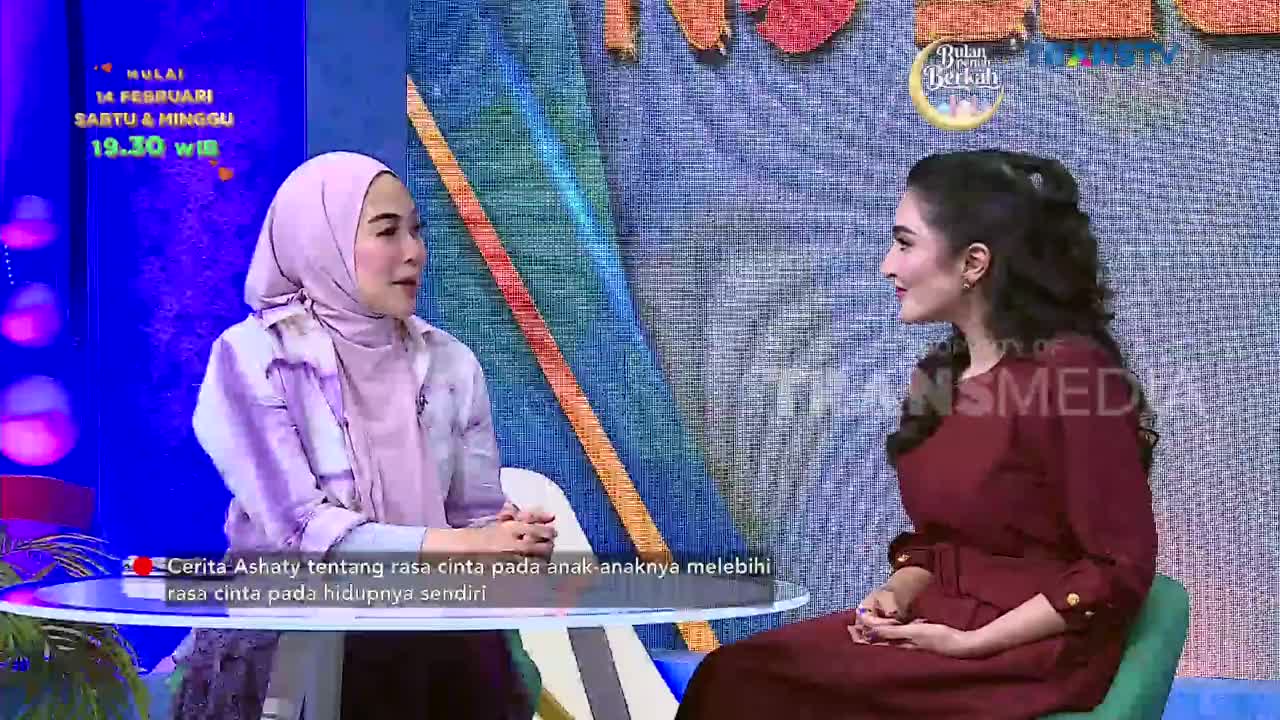 Two women are seated on a television set, one wearing a hijab and the other a maroon dress, engaged in conversation. The Trans TV logo is visible on the screen behind them.