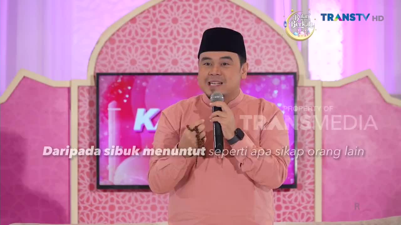 A man in a peach-colored shirt and black peci holds a microphone, speaking into it. He gestures with his free hand, a string of prayer beads visible. The Trans TV logo appears on the screen behind him.