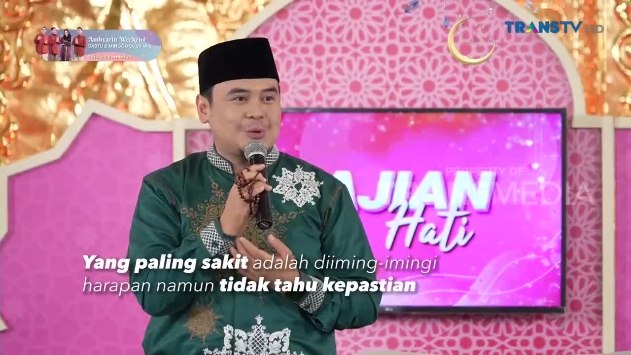 The man in the green shirt speaks into a microphone, his hand holding prayer beads. Behind him, a screen displays the Trans TV logo and the title "AJIAN Hati."