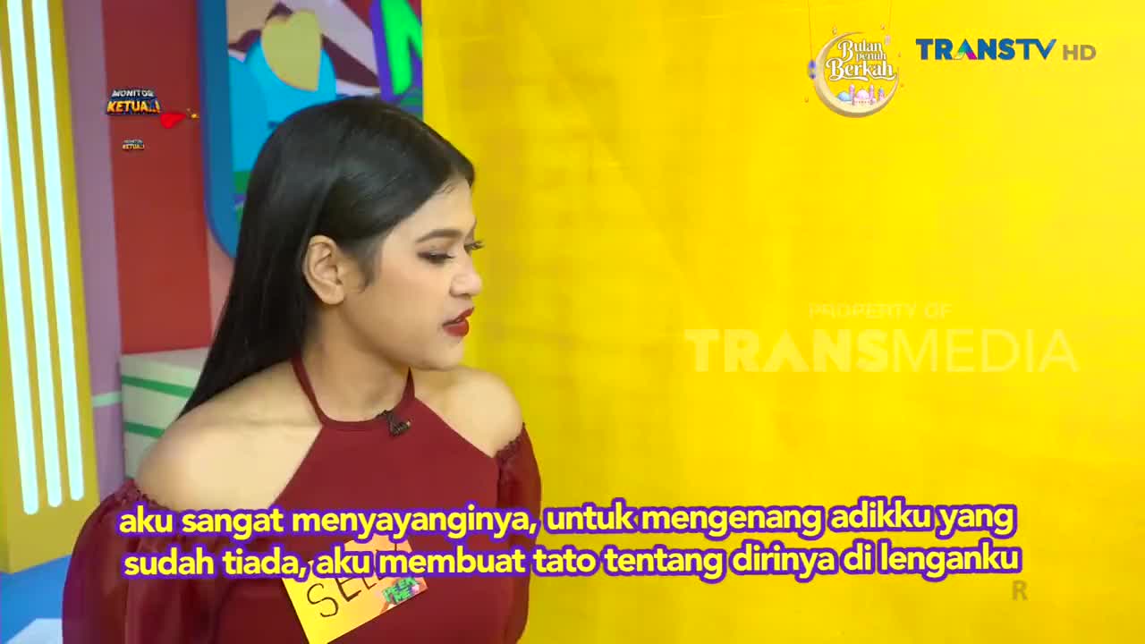 A young woman with long, dark hair speaks, her maroon top off-the-shoulder. The Trans TV logo appears on a bright yellow background behind her.