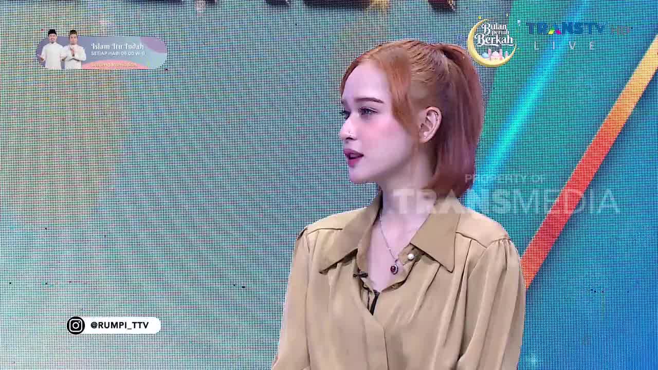 A woman with auburn hair pulled into a ponytail speaks on a Trans TV program. The background displays a grid pattern with a colorful diagonal stripe.