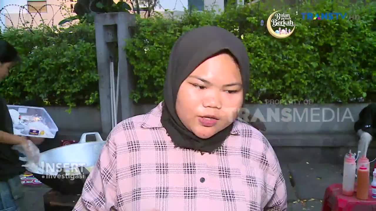 A young woman in a pink plaid shirt and hijab speaks to the camera, while behind her, someone prepares food in a large pot. The Trans TV logo is visible in the background, indicating this is a segment from Indonesia.