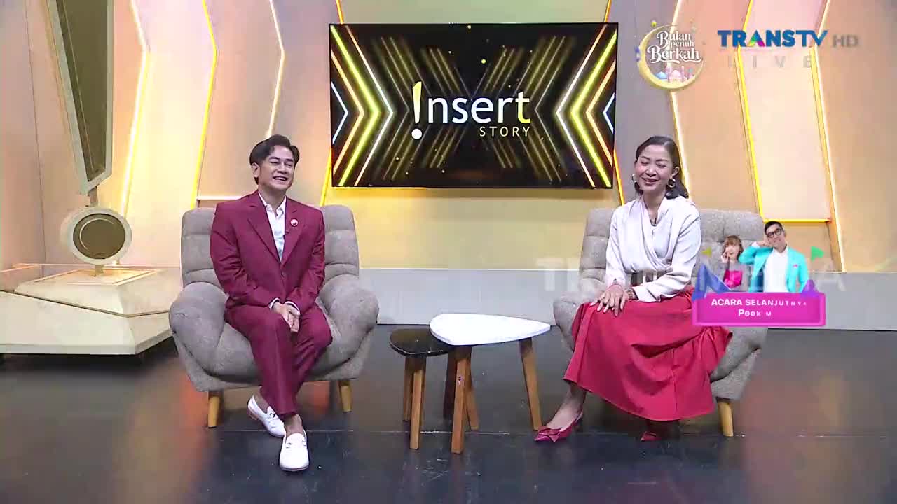 A man in a maroon suit and a woman in a red skirt sit in chairs on a studio set for Trans TV's "Insert Story." The screen behind them displays the show's logo, illuminated by bright, angled lights.
