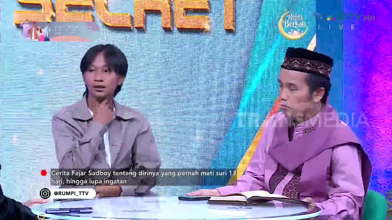 A young man in a tan jacket sits with his hand to his neck, listening intently. Across from him, a man in a purple robe and peci, with a book open before him, looks down. The Trans TV logo is visible on the screen behind them.