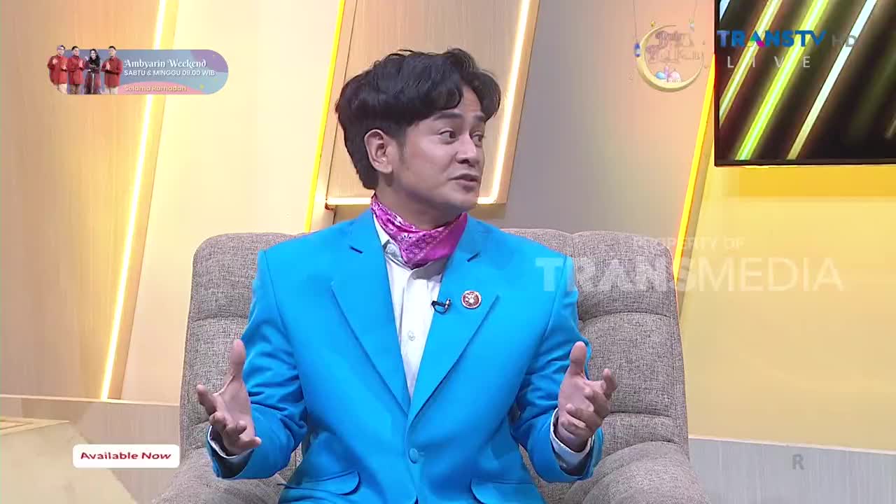 The man in the bright blue suit gestures with his hands as he speaks. A pink scarf peeks out from his collar, and the Trans TV logo is visible on the screen behind him.
