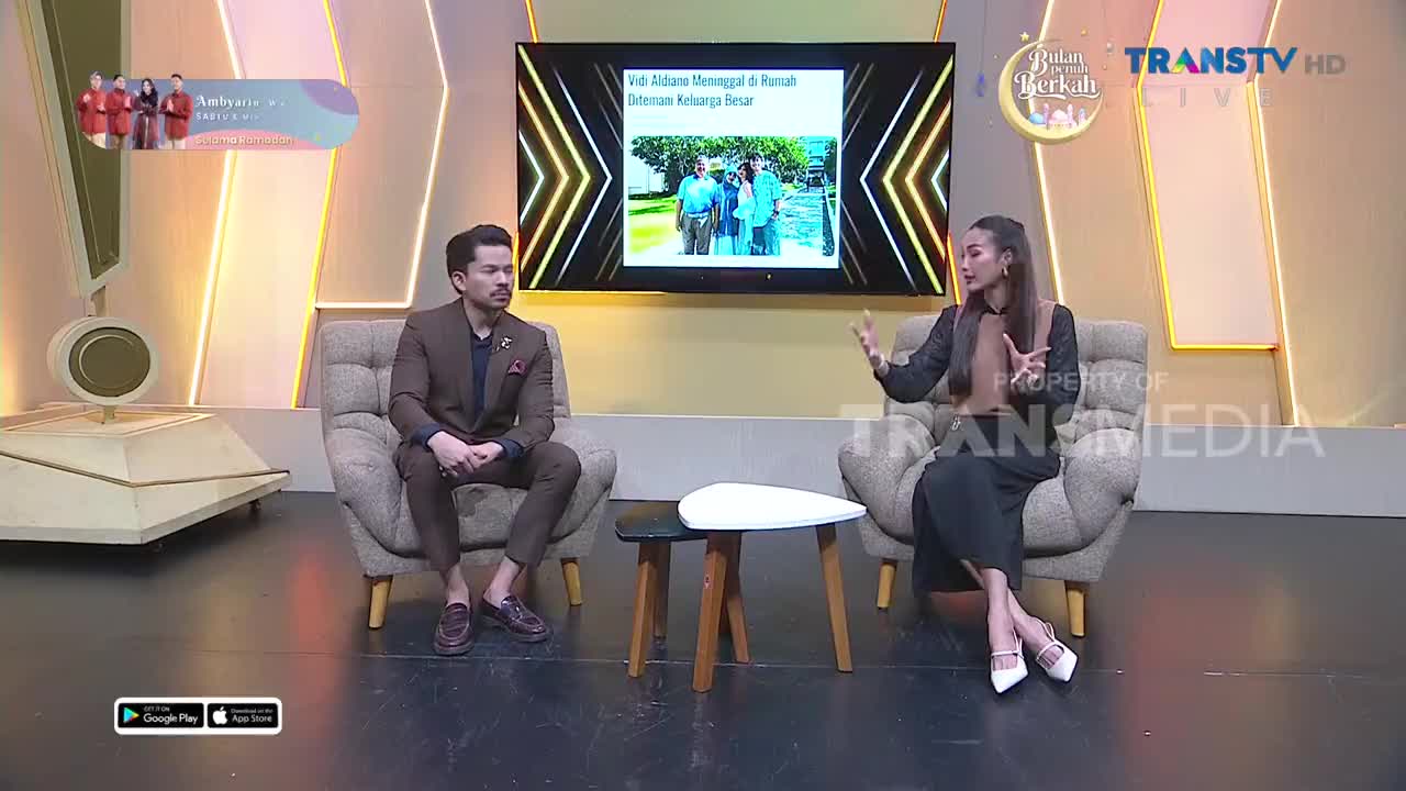 A man in a brown suit sits across from a woman in dark clothing, both on a TV show set for Trans TV. A large screen behind them displays a photo of people standing outdoors.