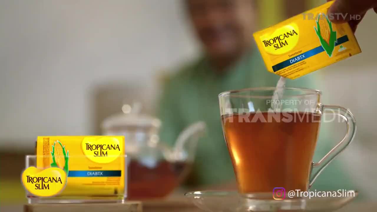 A hand tilts a yellow packet, pouring white sweetener into a clear glass of tea. Another packet of the same sweetener sits on display beside a teapot.
