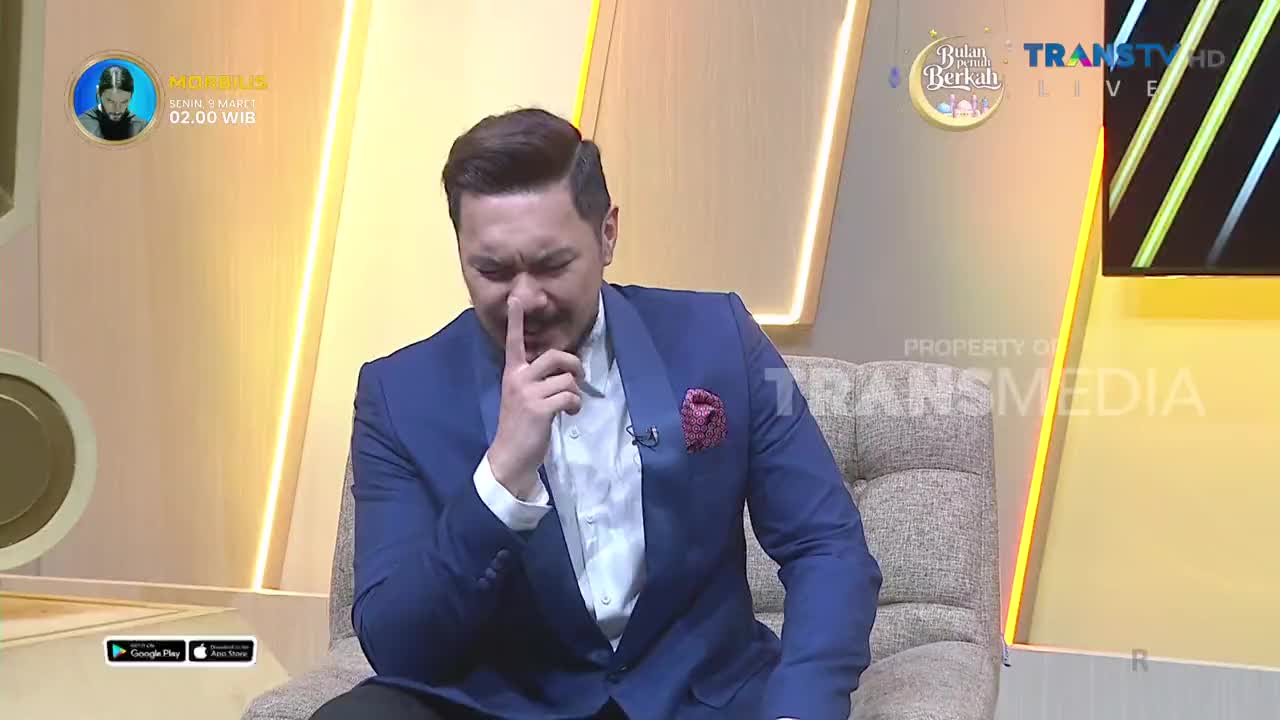 A man in a blue suit is sitting on a couch, holding his finger to his nose. He appears to be on a television set, with the Trans TV logo visible in the background.