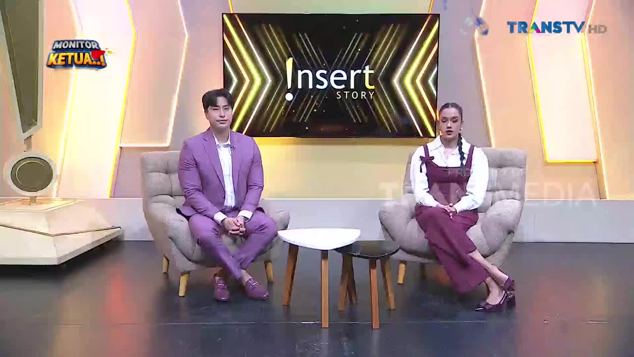 A man in a purple suit and a woman in a burgundy jumper sit in plush chairs on a brightly lit studio set. Behind them, a large screen displays the "Insert Story" logo, with "TRANS TV HD" visible in the upper right corner.