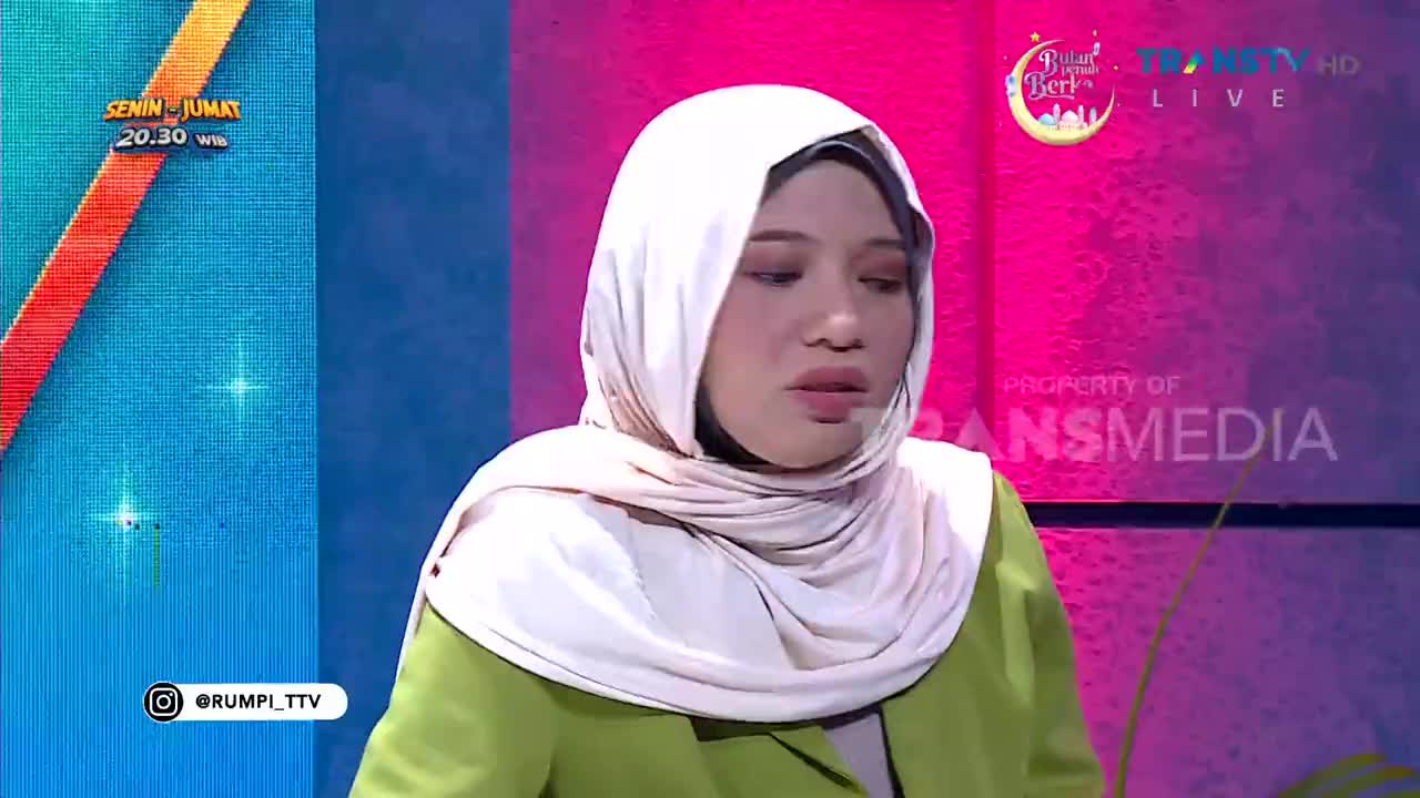 A woman in a pale hijab and a lime green jacket speaks on a brightly lit Trans TV set. The background displays a schedule for "Senin-Jumat 20.30 W."