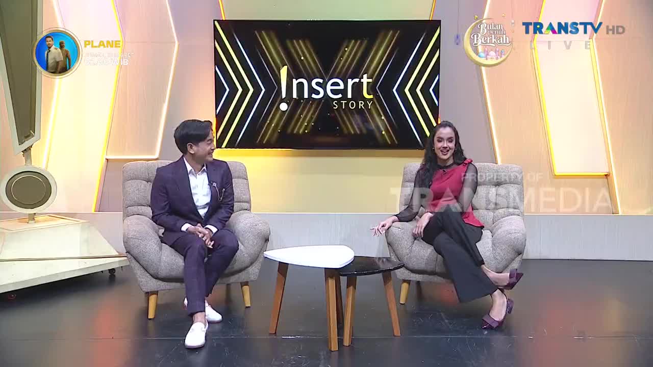 A man in a sharp suit and a woman in a stylish red top are seated on plush chairs, facing each other. Behind them, the "Insert Story" logo glows on a large screen, indicating a Trans TV production.