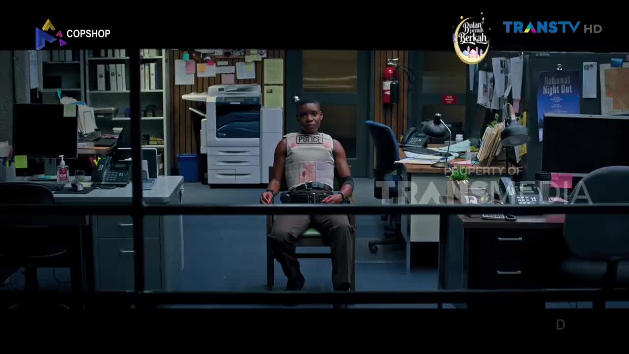 A woman in a police vest sits in an office, the Trans TV logo visible on screen. Papers and office equipment fill the room behind her.