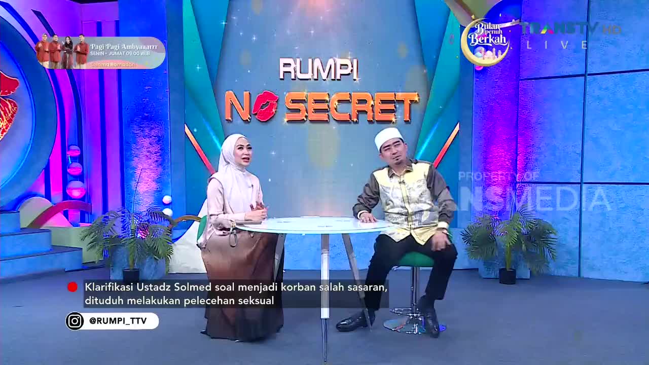 A woman in a hijab and a man wearing a peci sit across from each other at a small table on a brightly lit television set. The Trans TV logo is visible on the screen behind them, indicating this is a live broadcast in Indonesia.